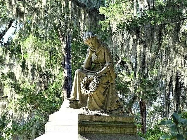 Statue einer sitzenden Frau mit einem Kranz auf einem moosigen Friedhof